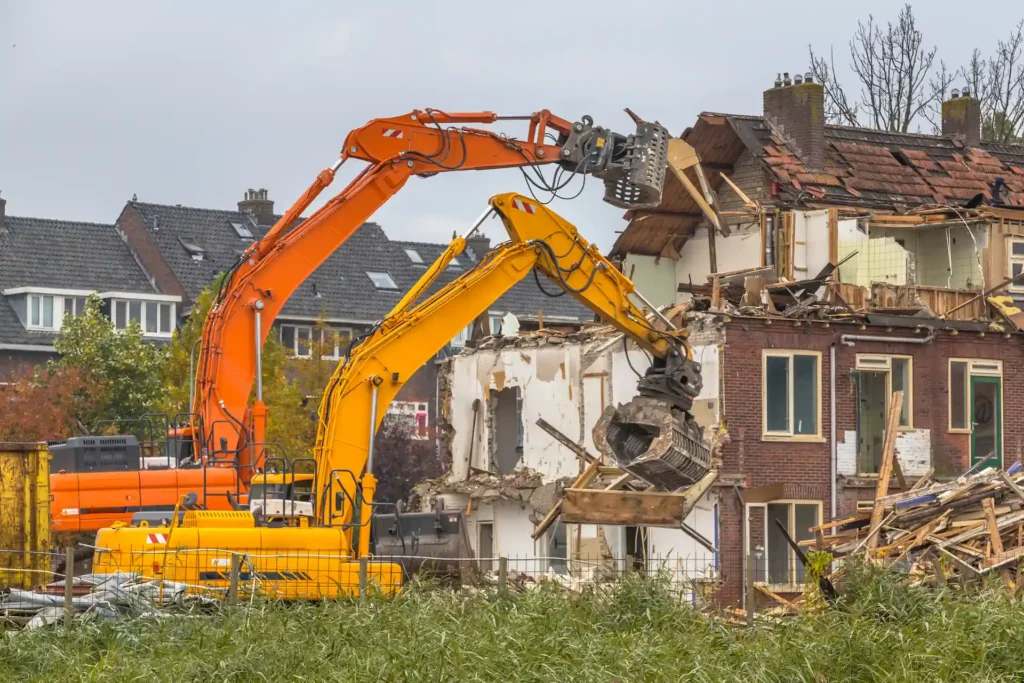 House Demolition - Large orange excavator with a mechanical claw demolishing a partially destroyed house.